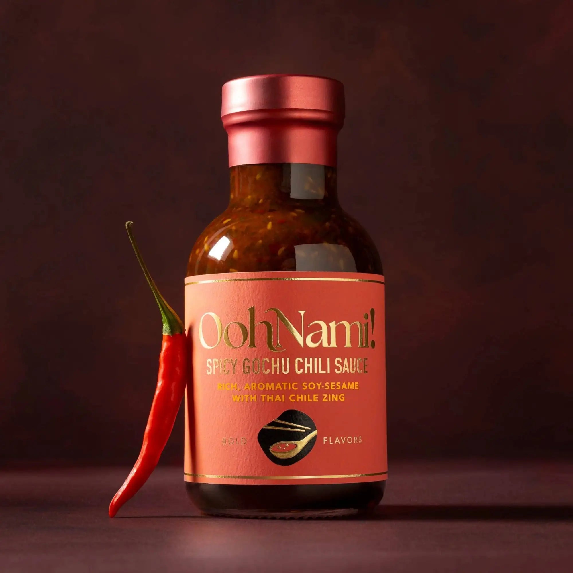 A bottle of ooh nami! Spicy gochu chili sauce with a reddish-pink label and metallic pink cap, containing rich aromatic soy-sesame sauce with thai chile zing.