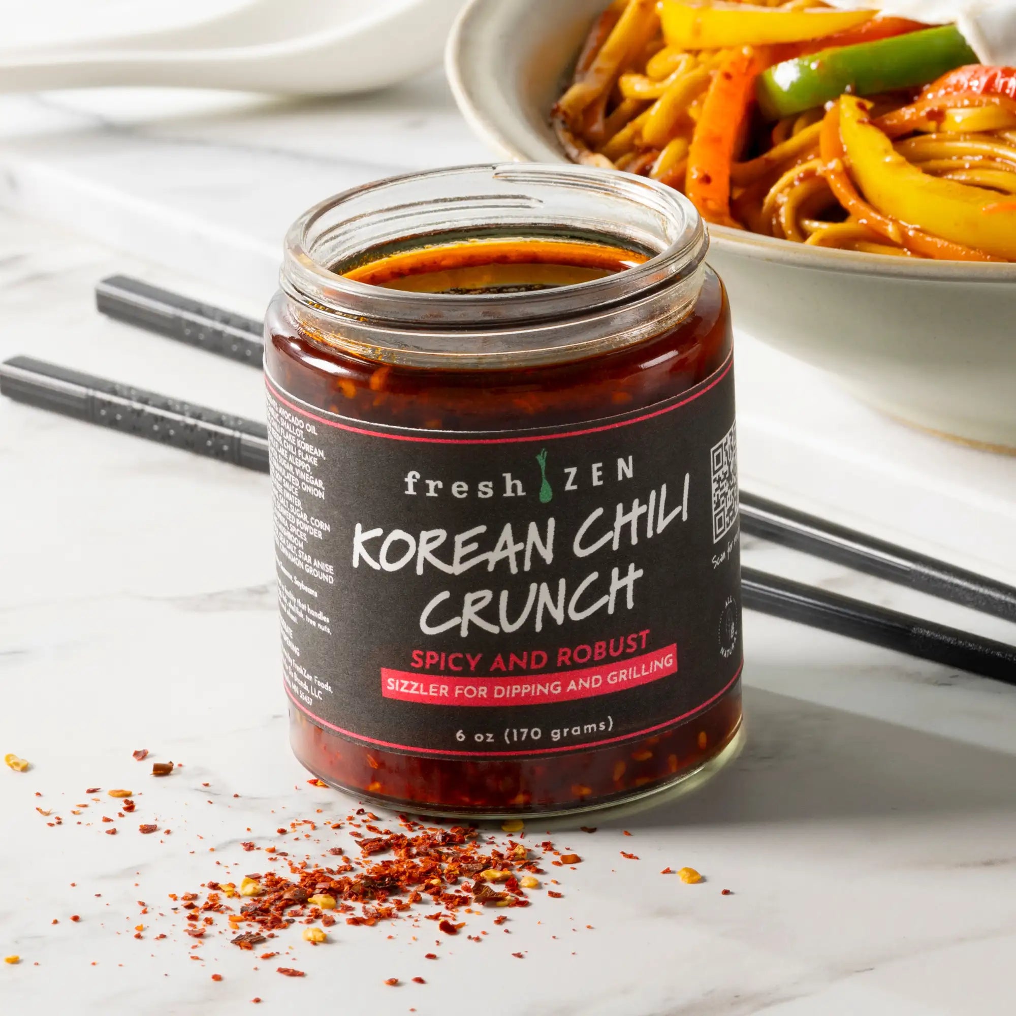 A glass jar of fresh zen korean chili crunch, featuring a black label with pink accents and red chili flakes scattered around its base.
