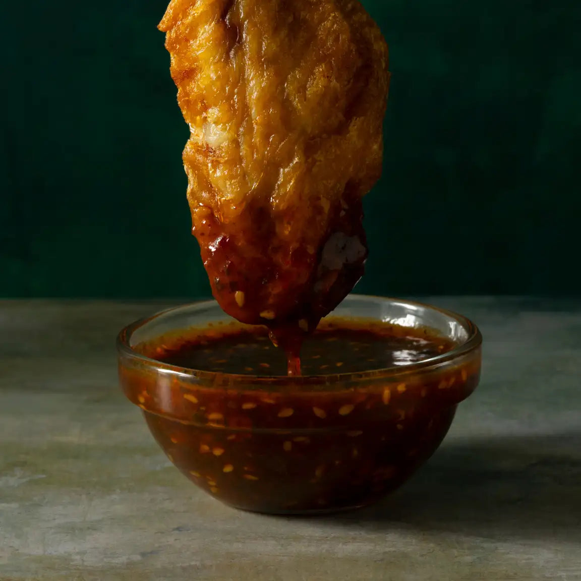 A golden-brown, crispy fried chicken wing dripping with a rich, red chili sauce from a clear glass bowl.