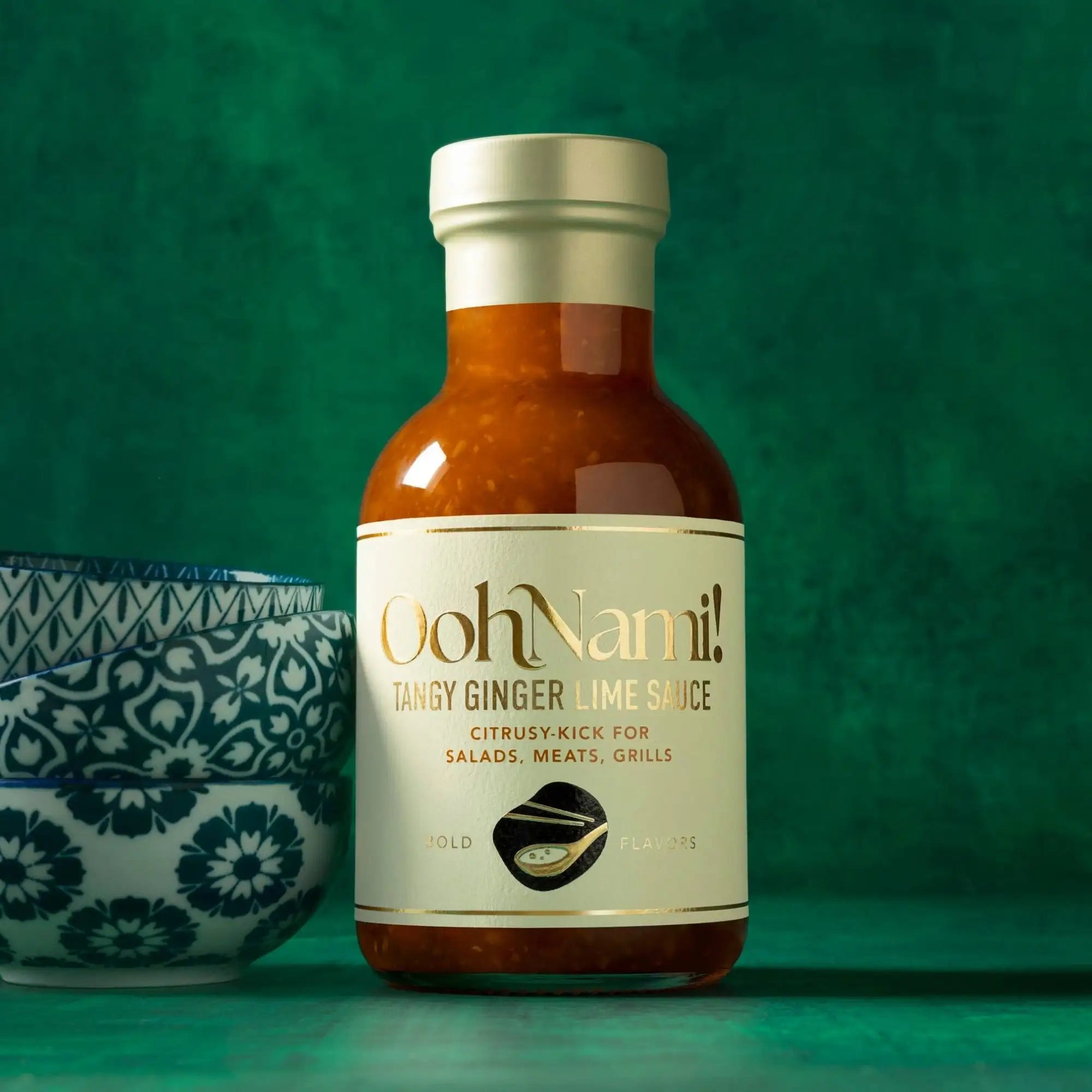 A sleek glass bottle of oohnami! Tangy ginger lime sauce with a metallic silver cap and cream-colored label featuring gold lettering, containing an amber-orange sauce.