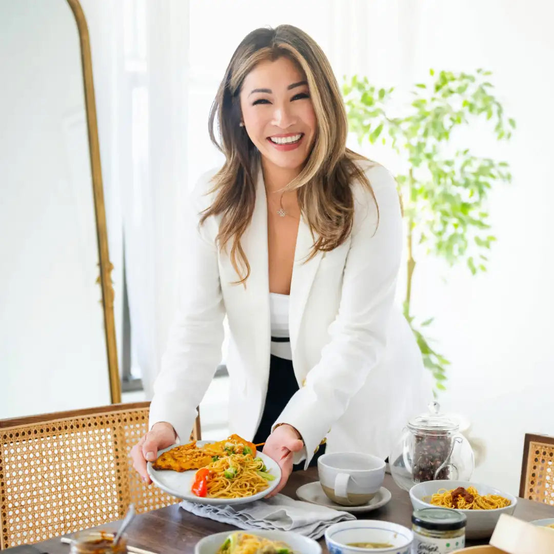 A woman in a white blazer holds a plate of colorful noodles and vegetables.