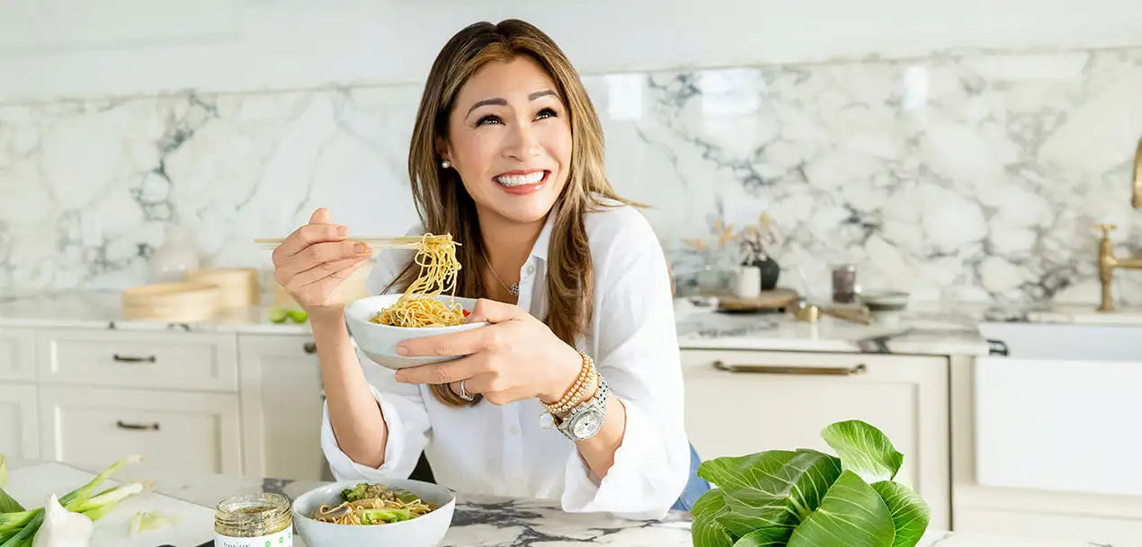 A woman in a white blouse holds a bowl of golden noodles with chopsticks, ready to eat.